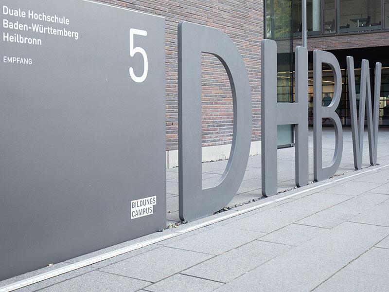 Die Abbildung zeigt den DHBW Schriftzug auf dem Bildungscampus Heilbronn. Er befindet sich vor dem Empfangsgebäude Bildungscampus 5, wie das Schild daneben betitelt.