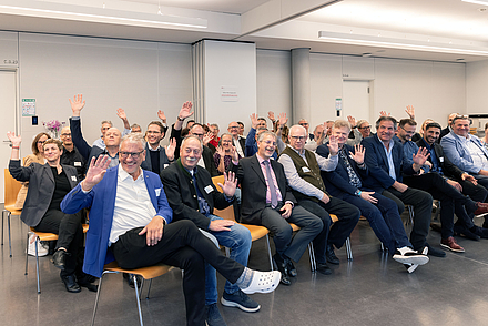 Die geehrten Dozentinnen und Dozenten sitzen in Stuhlreihen und winken in die Kamera.