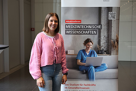 Das Bild zeigt die Studentin Alina Pfenninger vor dem Roll-up des Studiengangs Medinzintechnische Wissenschaften. Sie trägt ein rosa T-Shirt und Jeans.