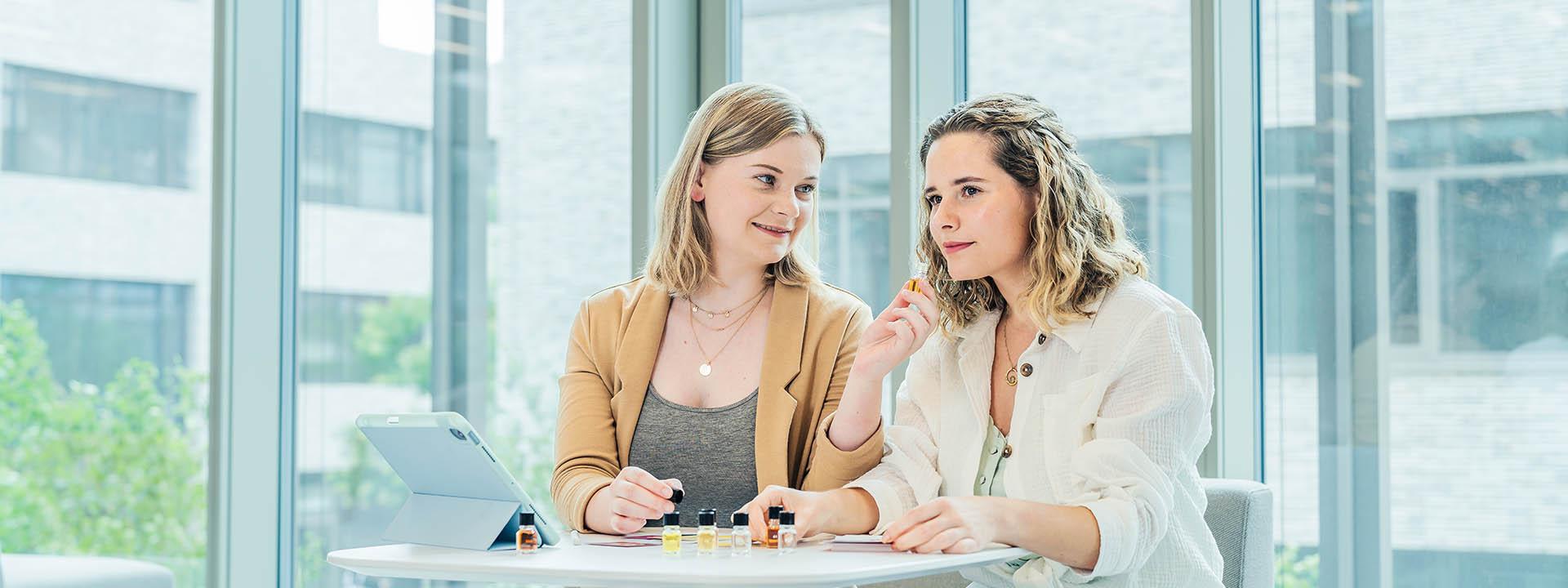 Geruchstest Das Foto zeigt zwei Frauen die an einem Tisch sitzen und kleine Glasfläschchen in der Hand halten. Auf dem Tisch stehen noch sechs weitere Glasfläschchen. Es sieht so aus, als würden sie einen Geruchstest durchführen.